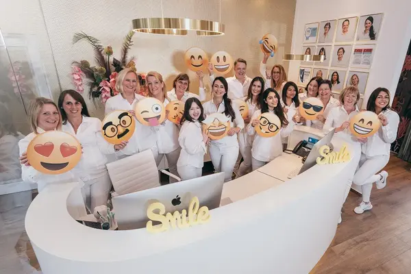 Team in weißer Kleidung lächelt an Rezeption einer Zahnarztpraxis und hält Emoji-Schilder.