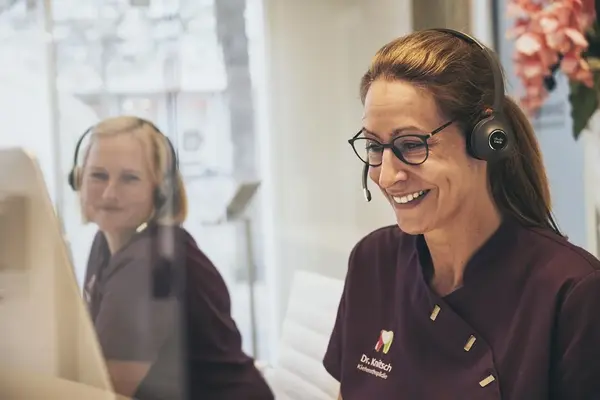 Zwei freundliche Zahnarztpraxis-Mitarbeiterinnen beraten Patienten am Empfang mit Headsets.