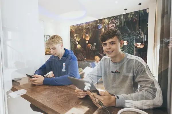 Zwei Jugendliche sitzen an einem Holztisch und nutzen Tablet und Smartphone in modernem Raum.