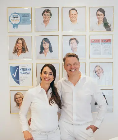 Zwei Zahnärzte im weißen Outfit stehen vor einer Wand mit Auszeichnungen und Teamfotos.